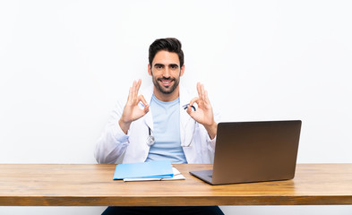 Young doctor man with his laptop over isolated wall showing an ok sign with fingers