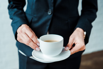 cropped view of woman holding cup of coffee and saucer in office