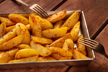 Potato wedges, oven baked, in a baking tray on a dark rustic wooden background, with two forks