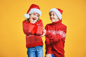 Happy boy in Santa hat giving Christmas gift box to excited girl in red sweater with deer on yellow background