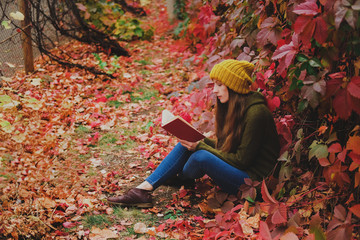Girl in mustard yellow knit hat and marsh green wool sweater sitting among colorful ivy in autumn and reading a paper book
