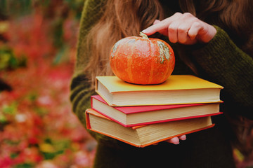 Female hands holding a stack of three vintage paper books and small pumpkin among colorful ivy in autumn.