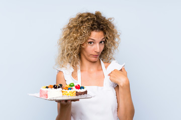 Young blonde woman with curly hair holding lots of different mini cakes with surprise facial expression