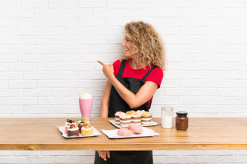 Young woman with lots of different mini cakes in a table pointing back with the index finger