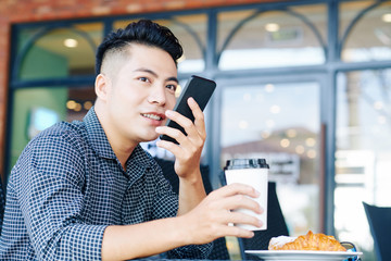 Positive handsome young VIetnamese man having cup of coffee and recodring voice message for...