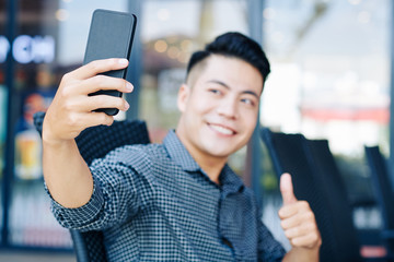 Handsome young Asian man showing thumbs-up when taking selfie on smartphone outdoors