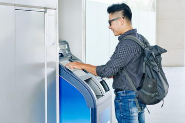 Smiling young Asian man entering pin code in ATM cash terminal to withdraw money