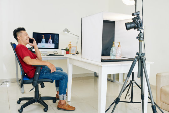Happy stock photographer sitting in chair and talking on phone after retouching photos of dispensers he made in his studio