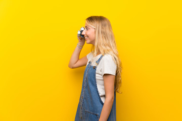 Blonde young woman over isolated yellow background holding a camera
