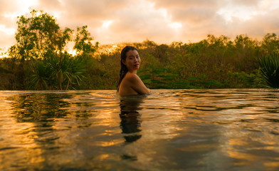 tropical holidays lifestyle portrait of young beautiful and happy Asian Japanese woman in bikini enjoying sunset at amazing jungle resort infinity pool swimming relaxed