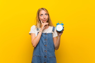 Blonde young woman over isolated yellow background holding vintage alarm clock