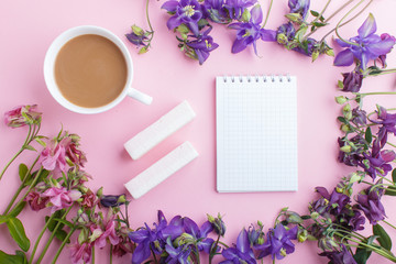 Pink and purple columbine flowers and a cup of coffee with notebook on pastel pink background.
