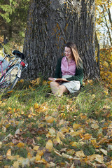 Young female with fall autumn leaves and red book in the hands in vintage old style clothes outdoors in the park or forest lean against an old tree and reading poetry, copy space