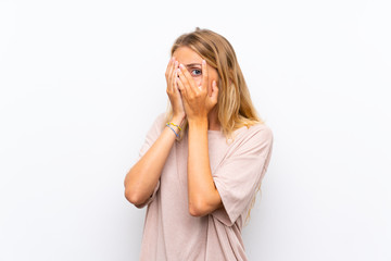 Blonde young woman over isolated white background covering eyes and looking through fingers