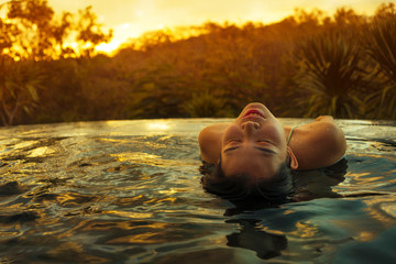 tropical holidays lifestyle portrait of young beautiful and happy Asian Chinese woman in bikini enjoying sunset at amazing jungle resort infinity pool swimming relaxed