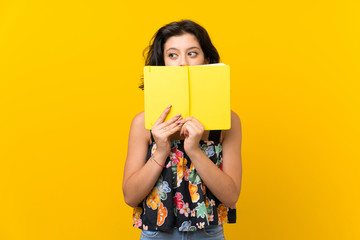 Young woman over isolated yellow background holding and reading a book