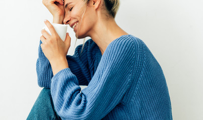 Happy gorgeous young woman dressed in cozy knitted blue sweater with a white mug with a tea in hand. Beautiful young woman drinking hot beverage at home. Smiling blonde female holding a coffee cup.