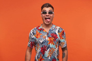 Portrait of funny pretty young male in flowered shirt and goggles having fun over orange background, looking joyfully to camera and showing tongue