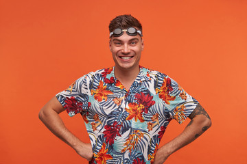 Happy attractive young swimmer with goggles on his forehead standing over orange background, looking at camera joyfully with hands on hips