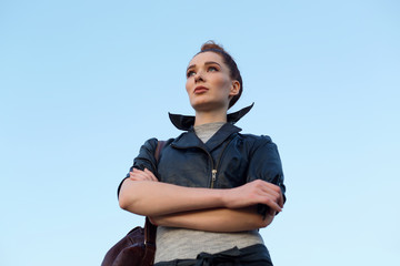 Beautiful red haired woman in a leather jacket standing on a background of blue sky.
