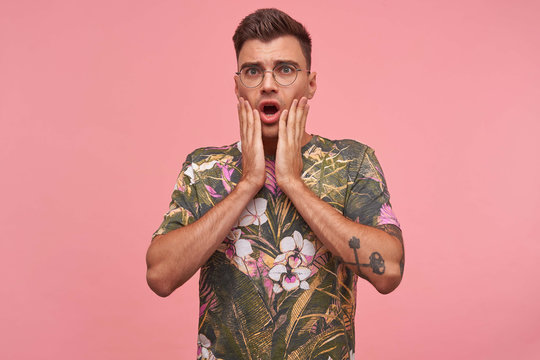 Handsome terrified male with covering mouth with hands, seeing something awful, wearing glasses and flowered t-shirt, isolated over pink background