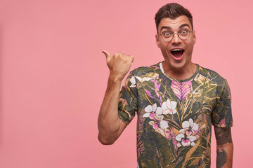 Indoor shot of excited short haired young man wearing glasses and flowered t-shirt, looking joyfully with surprised face expression, isolated over pink background