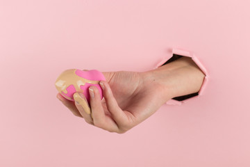 Girl hand holds a beauty blender in foundation, concealer from a hole in a pink background. Makeup artist concept, copy space.