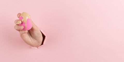Girl hand holds a beauty blender in foundation, concealer from a hole in a pink background. Makeup artist concept, copy space.