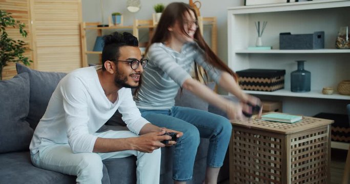 Girl and guy cute mixed race couple are enjoying video game at home using joysticks, woman is winning and celebrating success raising arms and laughing.