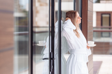 Pretty happy young woman in a white coat relaxes in a health spa hotel and enjoys a gorgeous view from her own terrace with a cup of coffee. Improvement and recovery concept