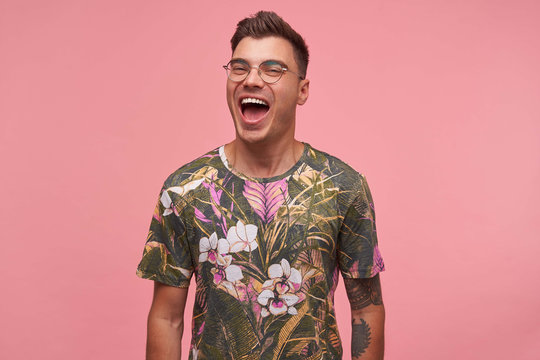 Indoor portrait of attractive young man with trendy haircut, looking happily and cheerfully to camera with hands down, isolated over pink background