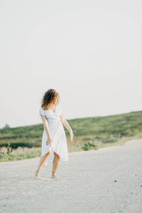 curly young woman dancing romantically in a blue dress on a sandy road at sunset.