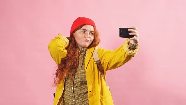 Young charming red-haired girl in tourist clothes takes a selfie on a pink background
