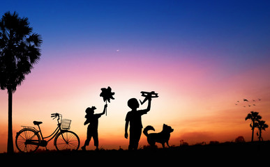 Fototapeta na wymiar Silhouete of kid dreams as pilot standing and holding airplane paper with friend running with wind turbine in sunset, imagination and freedom idea concept