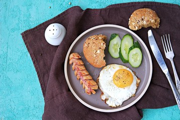 Breakfast, fried egg with sausage and slices of fresh cucumbers on a brown clay plate on a turquoise concrete background. Children's breakfasts. School breakfast concept. Top view, copy space.