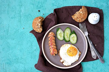 Breakfast, fried egg with sausage and slices of fresh cucumbers on a brown clay plate on a turquoise concrete background. Children's breakfasts. School breakfast concept. Top view, copy space.