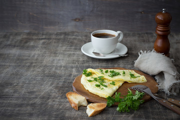 Morning breakfast of traditional French omelet with toasts, chopped parsley and white porcelain cup of coffee on a wooden board placed on a dark wooden table and dark wooden background, copy space
