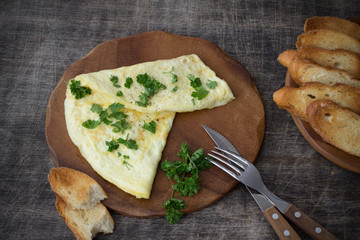 Morning breakfast of a traditional French omelet with toasts and butter, chopped parsley and a white porcelain cup of coffee on a wooden board placed on a dark wooden table, top view