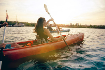 Happy young caucasian group of friends kayaking on river with sunset in the backgrounds. Having fun in leisure activity. Happy male and female model laughting on the kayak. Sport, relations concept.