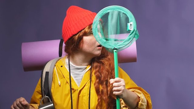 excited cheerful hiker wearing care mat, backpack, yellow coat, binoculars on neck holding butterfly net, close up photo. isolated blue background, studio shot, facial expression, reaction