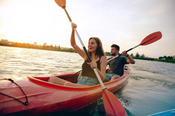Confident young caucasian couple kayaking on river together with sunset in the backgrounds. Having fun in leisure activity. Romantic and happy woman and man on the kayak. Sport, relations concept. - obrazy, fototapety, plakaty