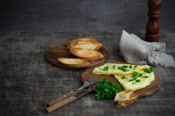 Morning breakfast of a traditional French omelet with toasts and butter, chopped parsley and a white porcelain cup of coffee on a wooden board placed on a dark wooden table, top view