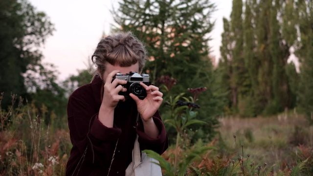 Trendy hipster girl with dreads taking pictures of plants in the garden with a vintage film camera at cold summer morning