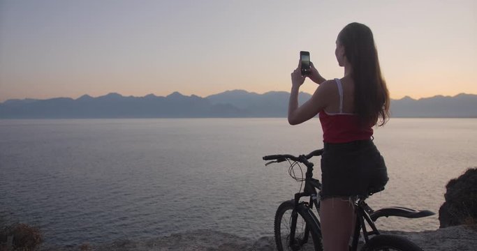Pretty lady on cliff with bicycle and making photo of sunset over sea with smartphone.
