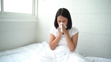 Flu.Rhinitis, cold, sickness, allergy concept. Young Asian woman got nose allergy, flu sneezing nose sitting at bed in bedroom,female. Brunette sneezing in a tissue