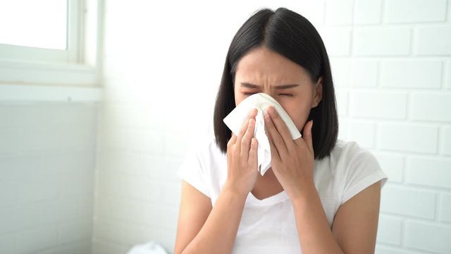 Flu.Rhinitis, cold, sickness, allergy concept. Young Asian woman got nose allergy, flu sneezing nose sitting at bed in bedroom,female. Brunette sneezing in a tissue