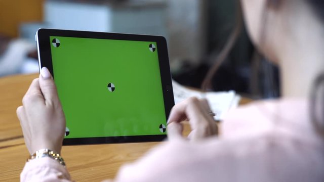View from the shoulder of young woman in pink shirt sitting at the table and sliding on her tablet at the chroma key green screen. Stock footage. Chroma key screen for placement of your own content.