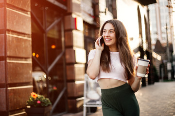 Smiling woman using phone and drink coffee on the street in summer day.