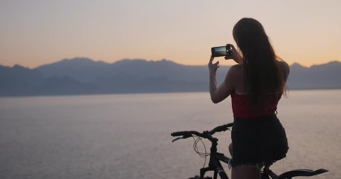 Pretty lady on cliff with bicycle and making photo of sunset over sea with smartphone.