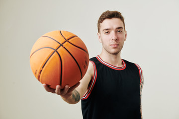 Handsome young serious Caucasian basketball player outstretching arm with ball and looking at camera
